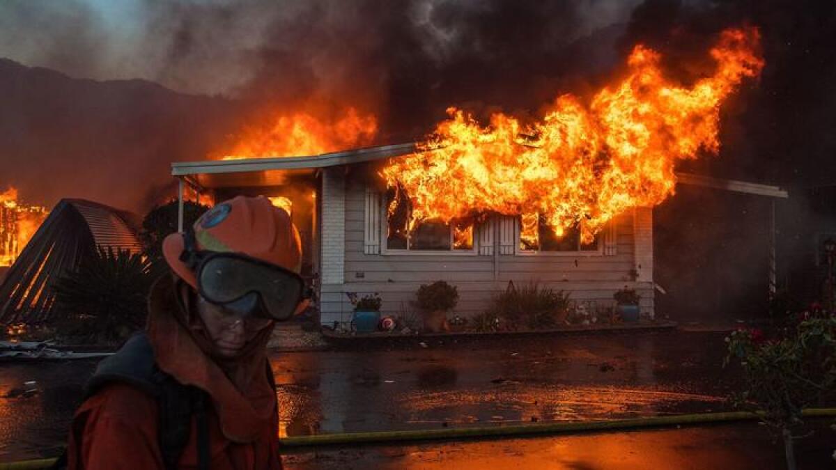 A firefighter in full protective gear stands in the foreground, silhouetted against the glow of a burning house, battling intense flames and smoke during a California wildfire.